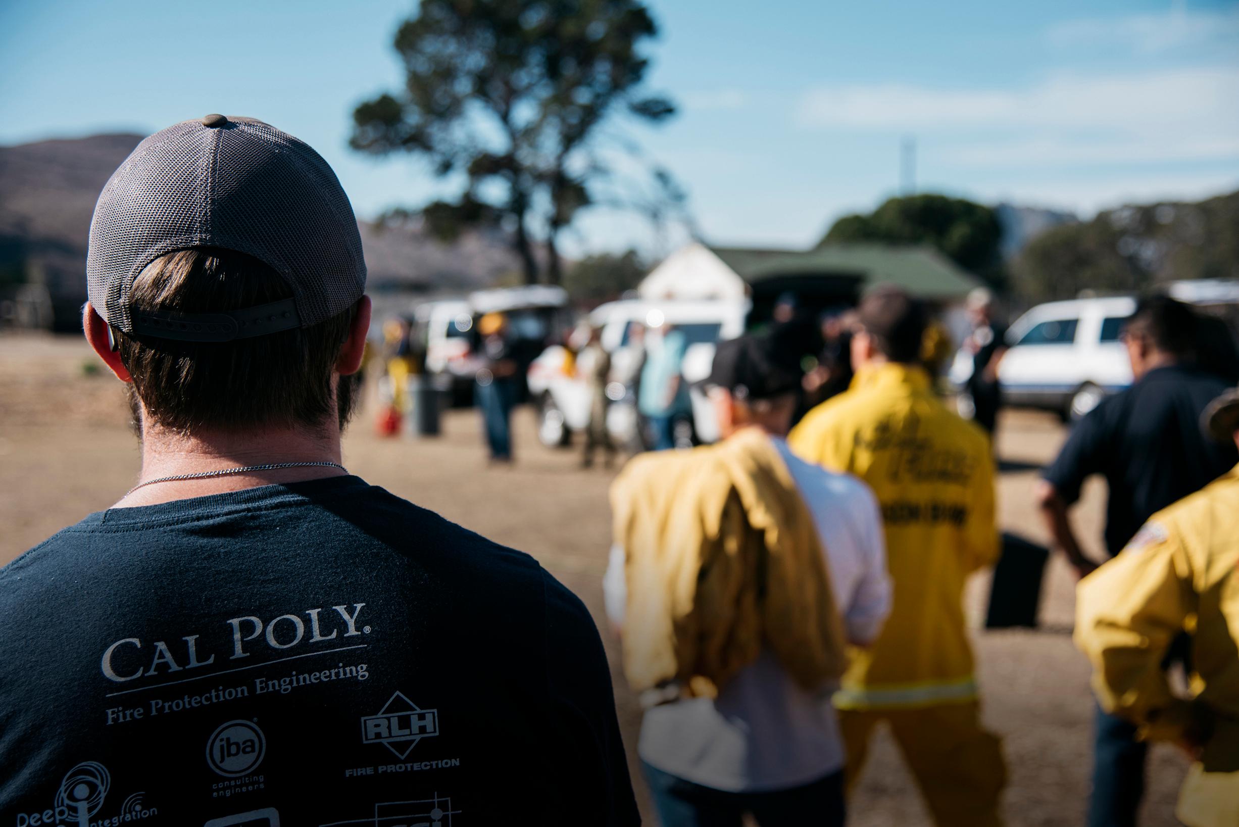 Person wearing a "Cal Poly Fire Protection Engineering" shirt watching a group of people, some in yellow fire gear, outdoors.