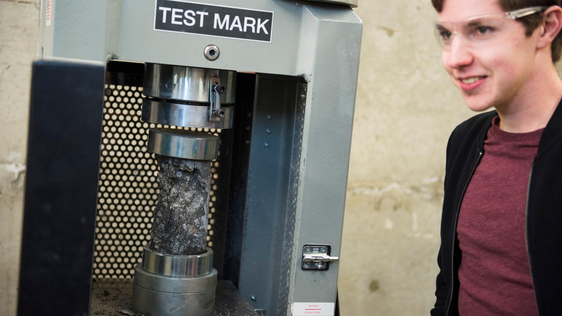 Student observes a concrete cylinder being compressed in a materials testing machine during an engineering lab.