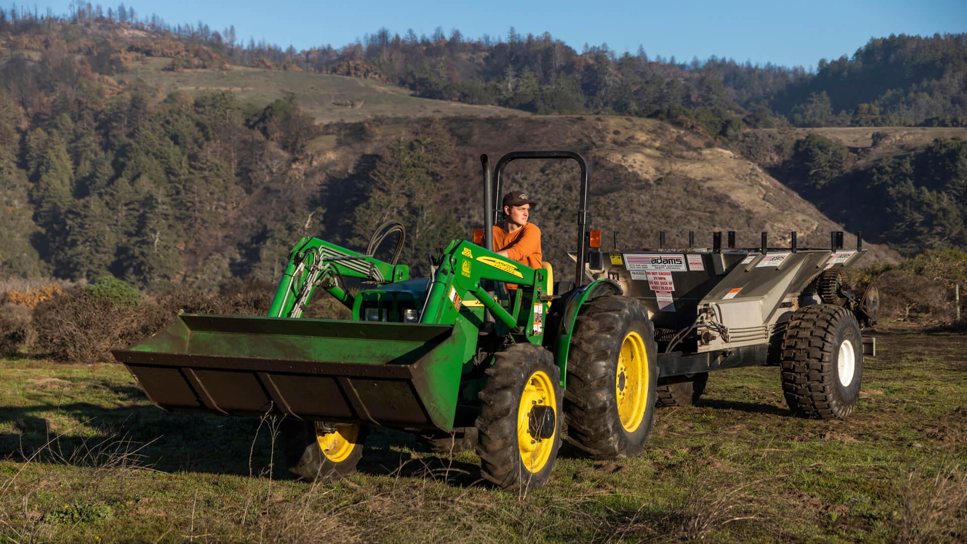 Student drives a green tractor pulling agricultural equipment across a field in a bioresource and agricultural systems lab.