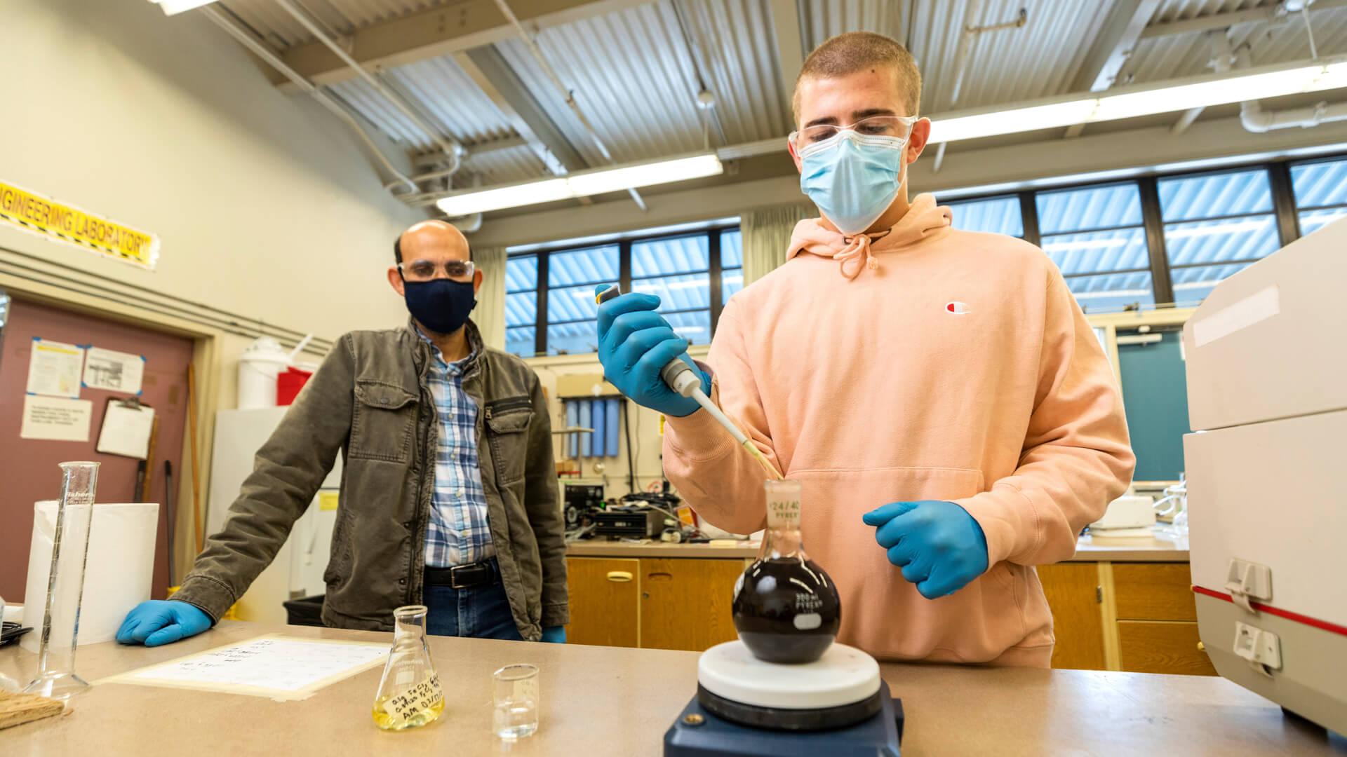 Student uses a pipette to transfer liquid into a flask while an instructor observes in a laboratory setting.