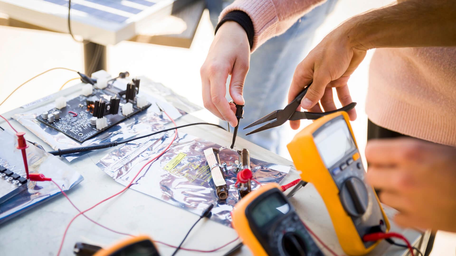 Students use pliers and multimeters to test and assemble electronic components on a workbench.