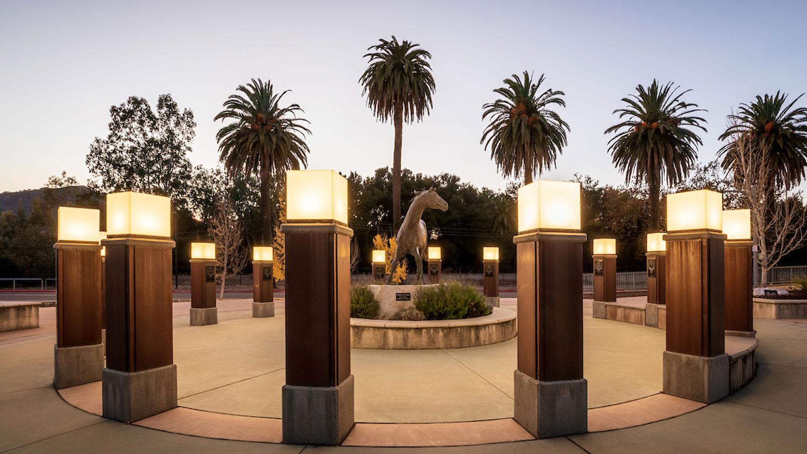 Campus plaza at dusk featuring illuminated columns and a horse statue surrounded by palm trees.