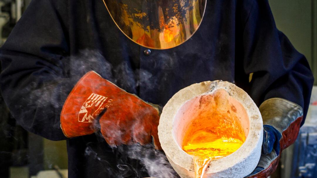 Person in protective gear pouring molten metal from a crucible in a manufacturing lab.