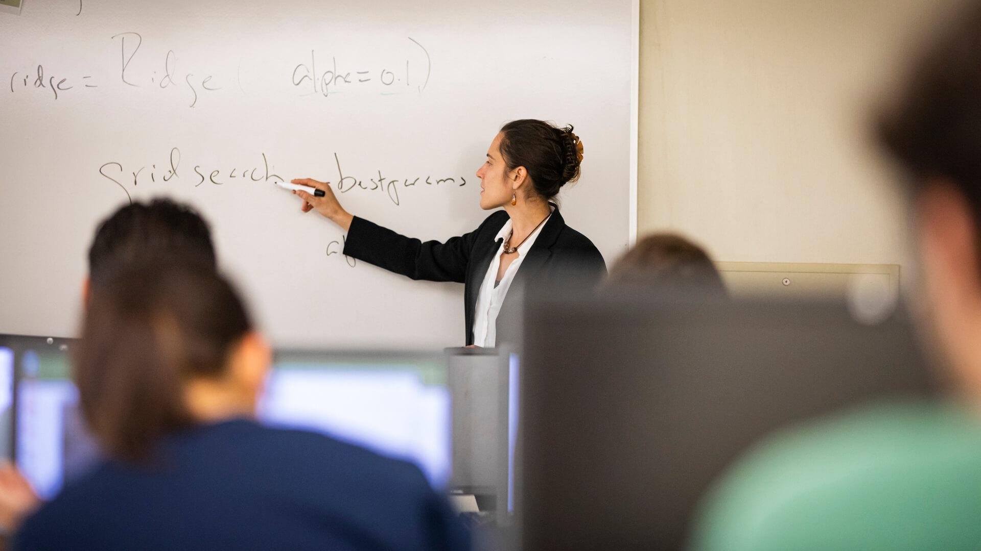 Professor writing equations on a whiteboard while teaching students in a computer lab.