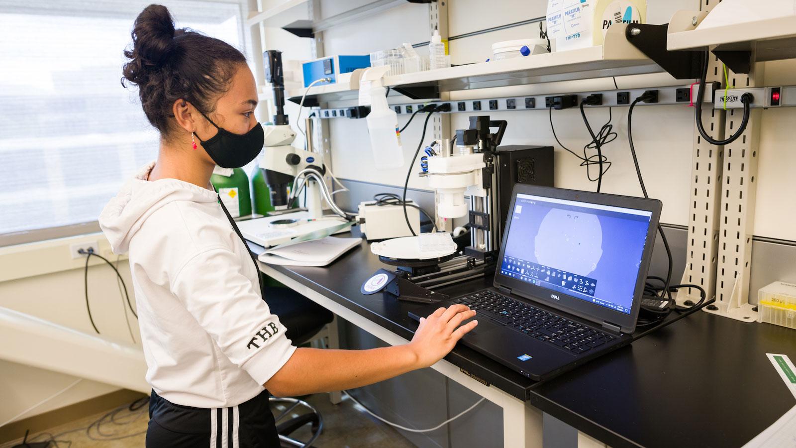Student operating laboratory imaging equipment while viewing results on a laptop.