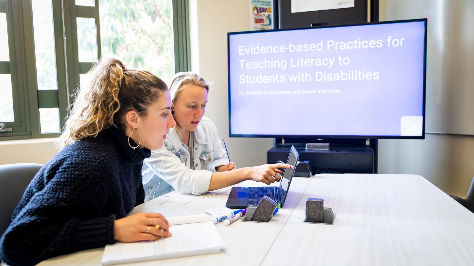 Students reviewing instructional materials on a laptop with a presentation about literacy for students with disabilities displayed on a screen.