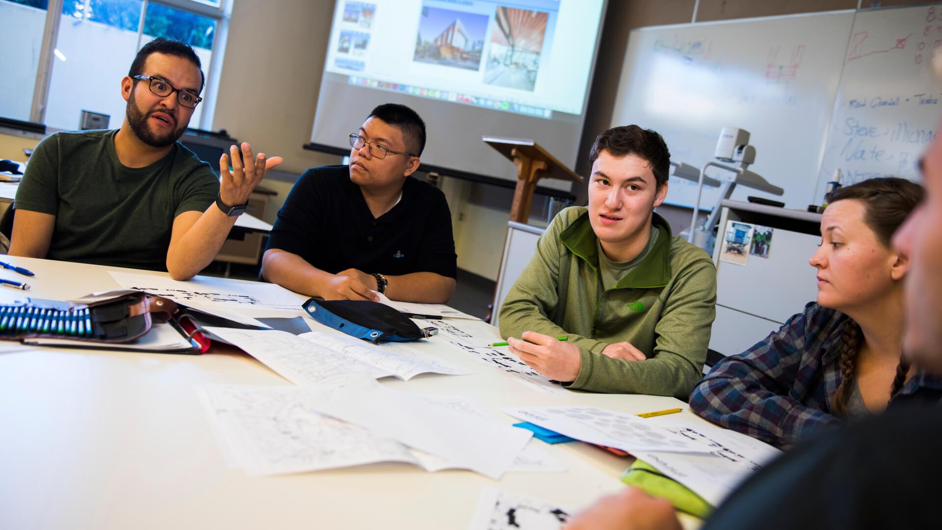 Group of students gathered around a table reviewing maps and planning documents during a classroom discussion.