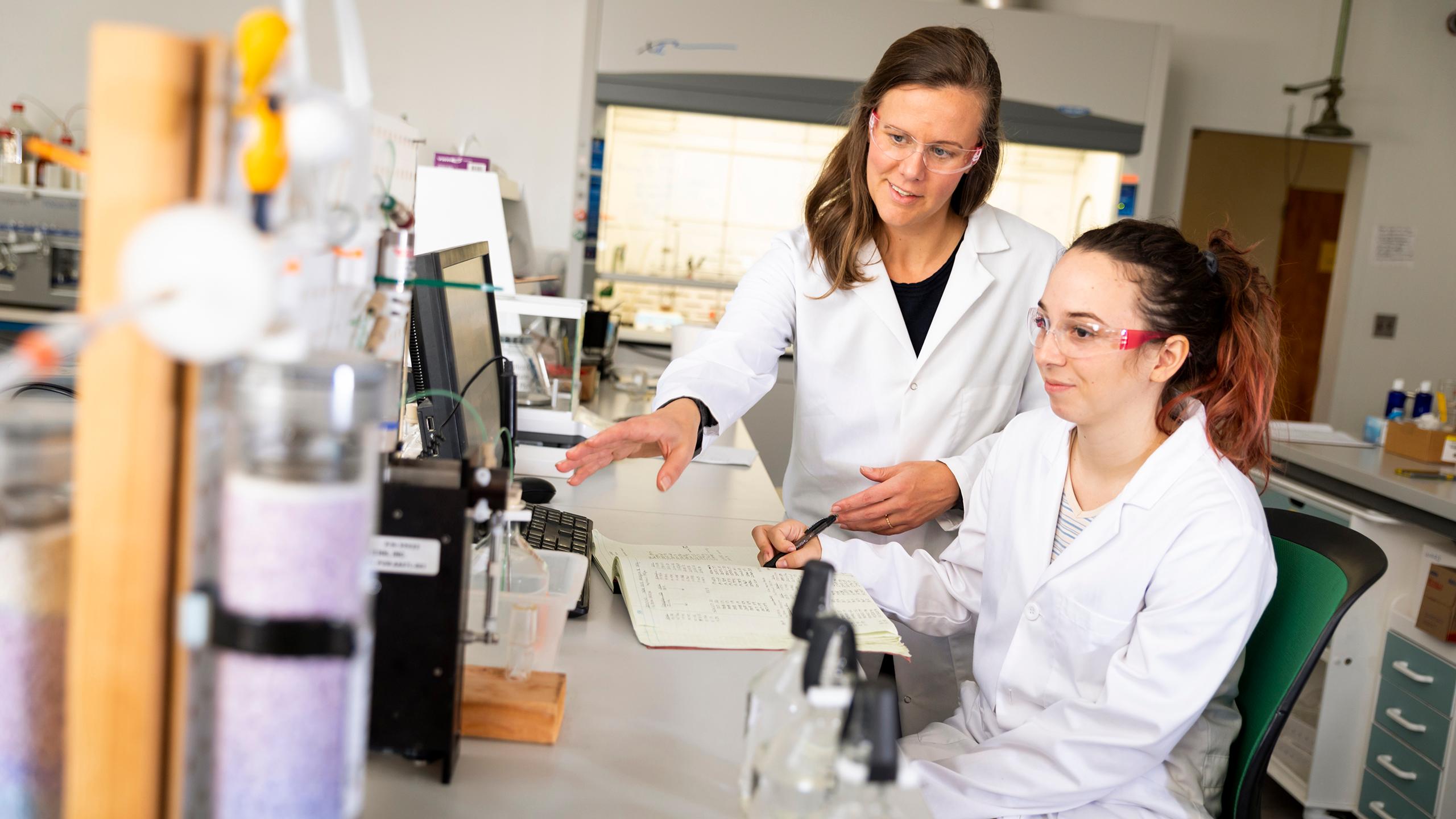 Two women in lab coats and safety glasses working at a computer in a laboratory with scientific equipment.