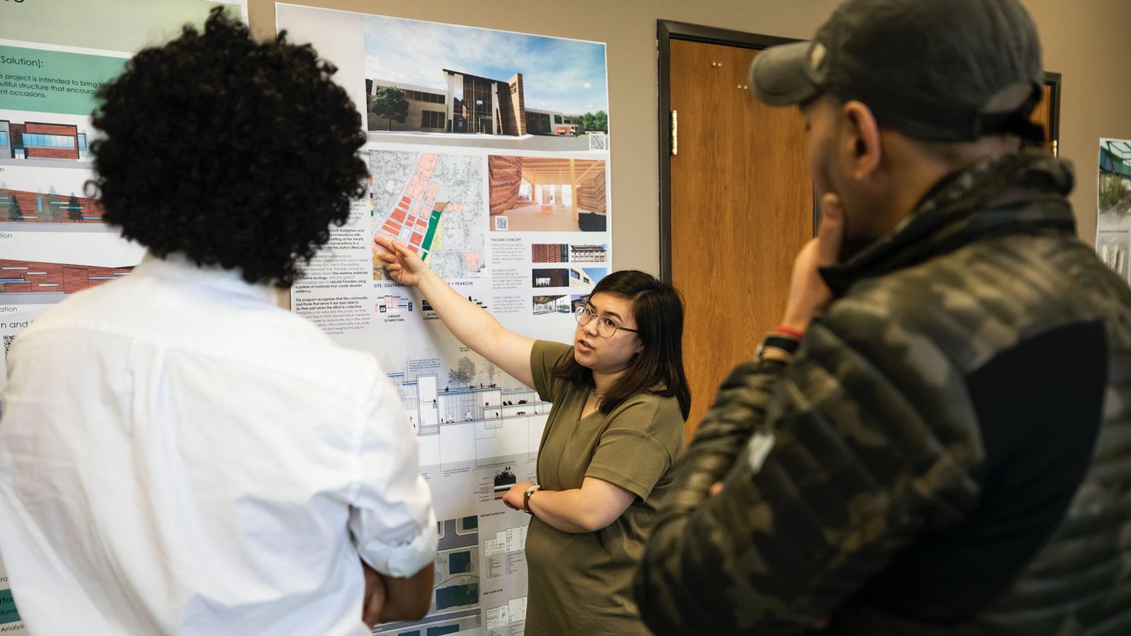 Woman explaining architectural project posters to two men in an indoor setting.