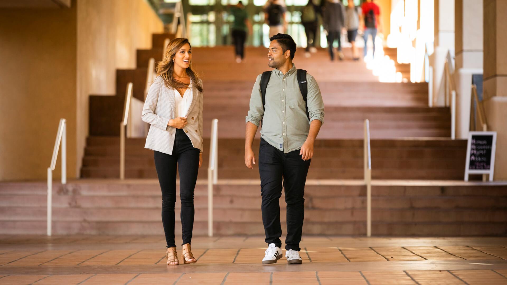 Two college students walk and talk on campus steps, carrying backpacks between classes.