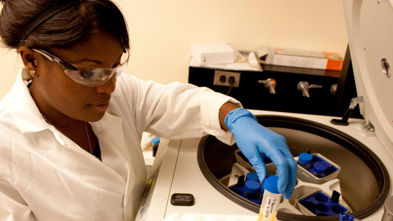 Scientist in lab coat and safety glasses placing sample containers into a laboratory centrifuge.