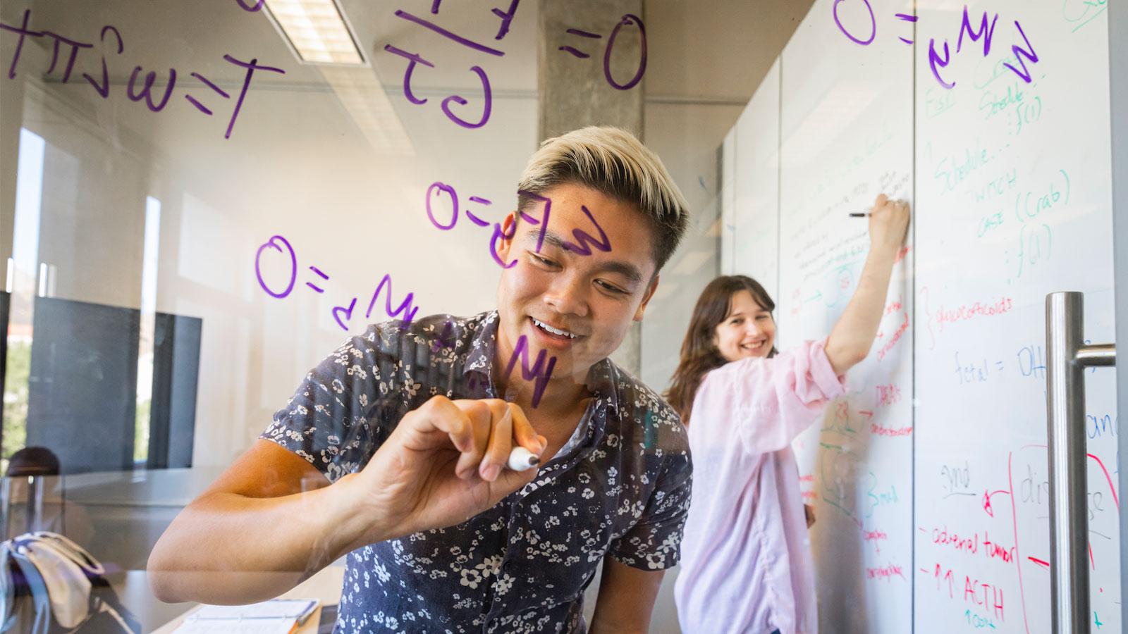 Students writing equations on a glass board while collaborating in a study space.