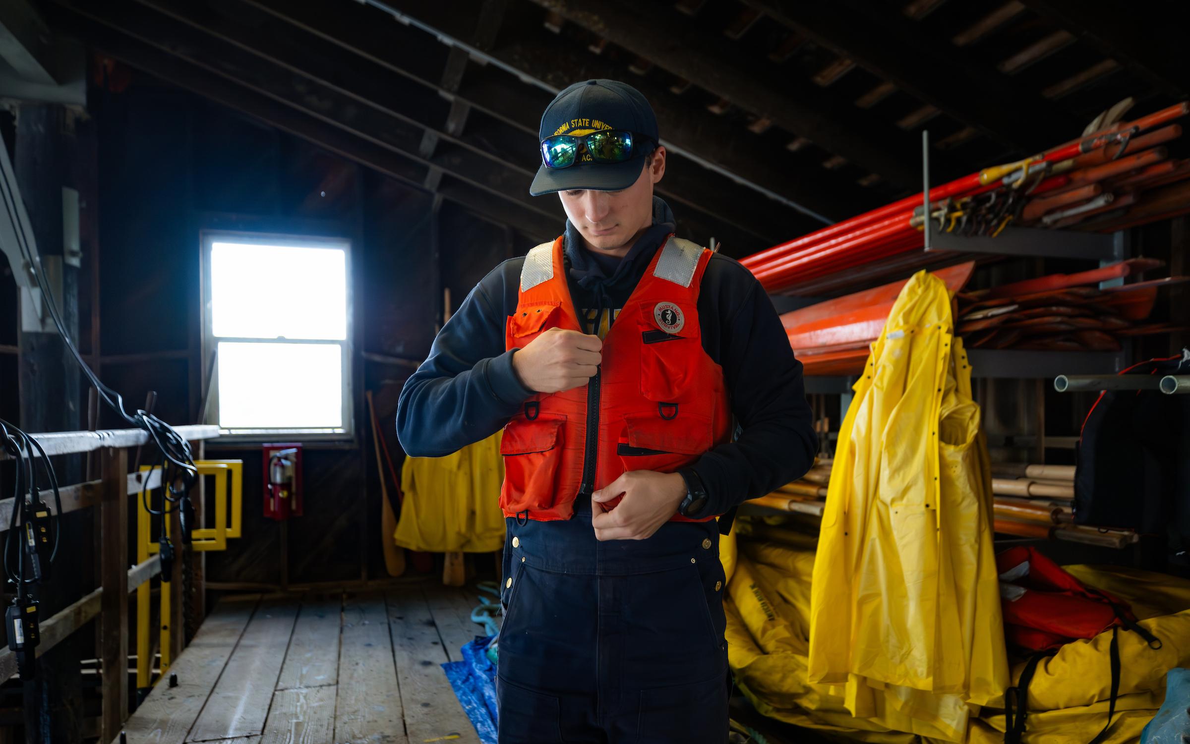 Person wearing a black cap and sunglasses putting on an orange life jacket in a wooden storage room with yellow gear.