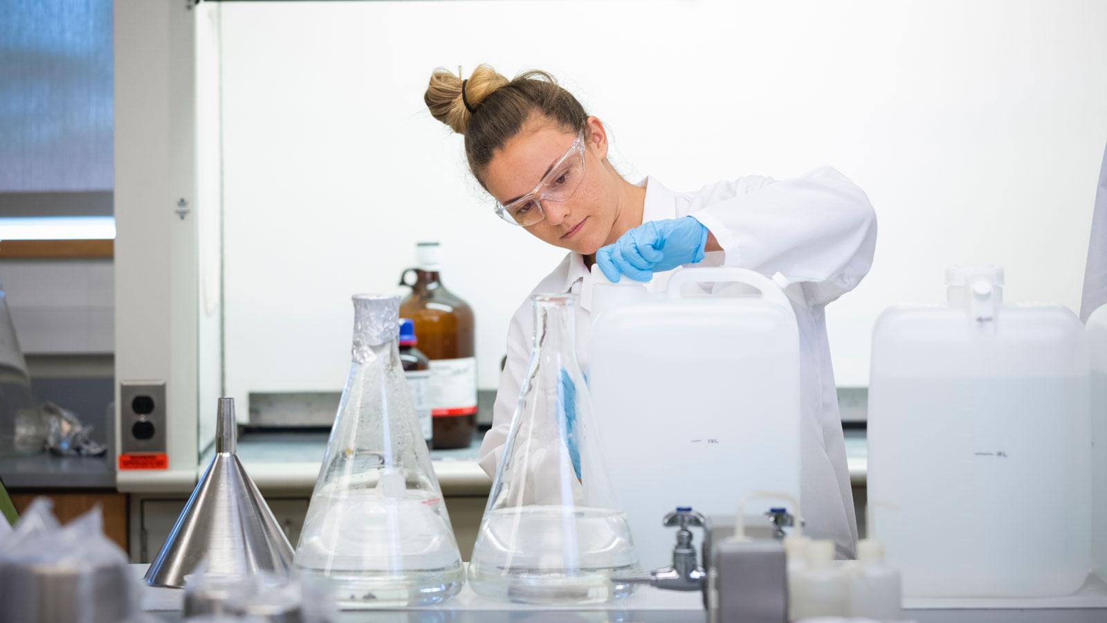 Woman wearing lab coat, gloves, and safety glasses pouring liquid from a large container in a laboratory.