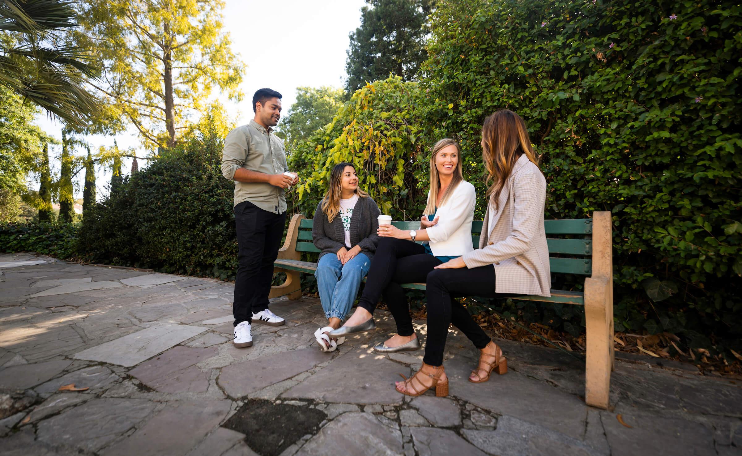 Four people socializing outdoors by a green bench, two sitting and two standing, surrounded by lush greenery.
